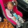 toddler, child, car_seat, car_interior, pink, curly_hair, white_bow, pacifier, blue_cup, butterfly_pants, jacket, orange, clothing, seatbelt, playful_expression, vehicle, window, door, seat, candid