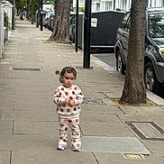 Arianna joined the competition — help win amazing prizes! toddler, child, sidewalk, street, trees, cars, urban, buildings, clothing, footwear, person, hairclip, pavement, daytime, outdoor, curious, standing, matching_outfit, casual, city