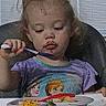 toddler, child, high_chair, spoon, plate, food, curly_hair, purple_shirt, frozen_characters, indoor, eating, meal, window_blinds, table, concentration, young_child, person, dining, seat, messy_face