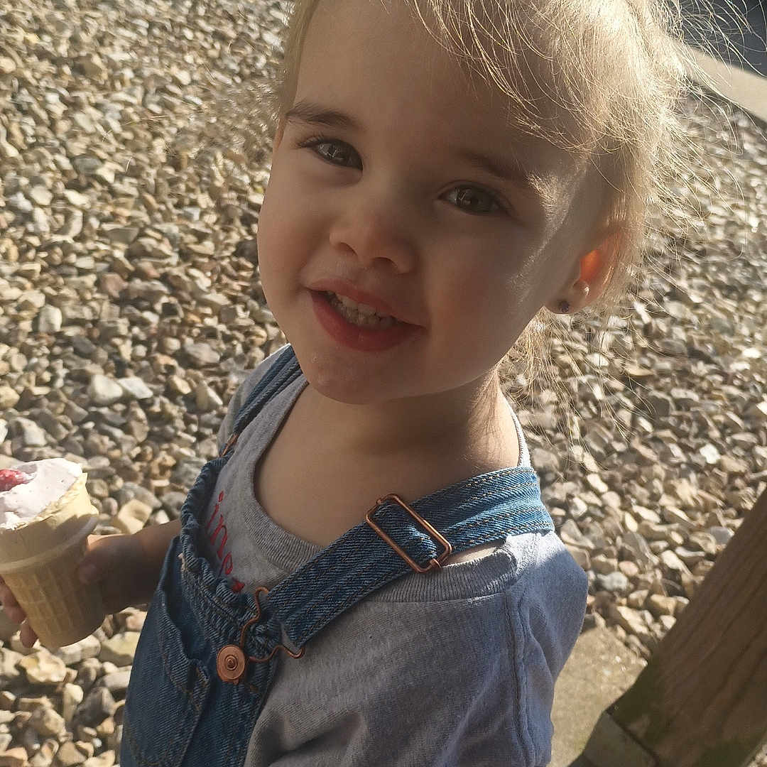 Linetta is registered to the contest to win money with this photo: barefoot, casual, child, cute, denim_overalls, face, grass, gravel, gray_shirt, hair, happy, ice_cream, outdoor, person, playful, portrait, smiling, summer, sunlight, toddler