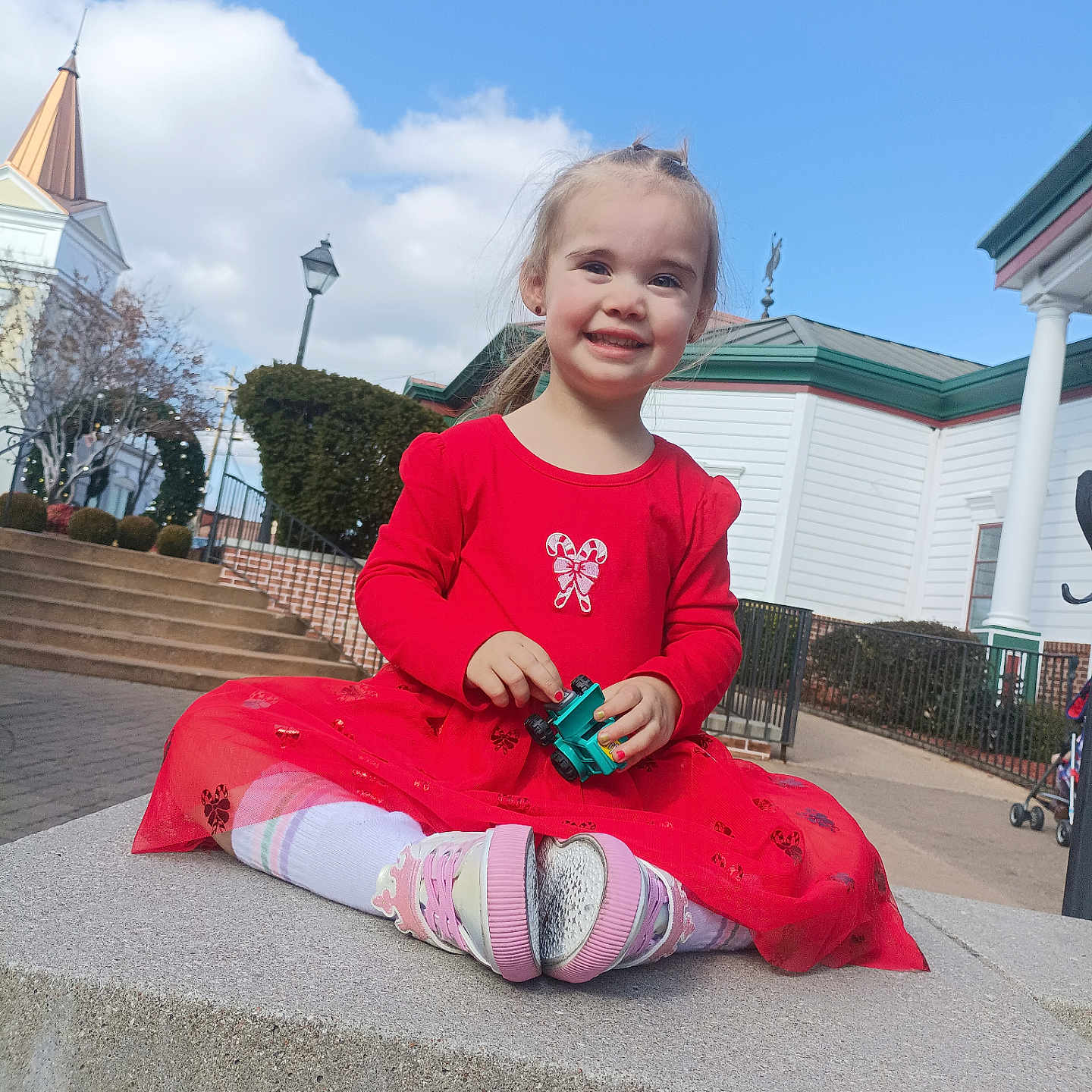Linetta is registered to the contest to win money with this photo: child, girl, red_dress, toy_car, smile, outdoor, blue_sky, clouds, concrete, steps, building, lamp_post, fence, shoes, tights, hair_tie, playful, happy, portrait, daylight