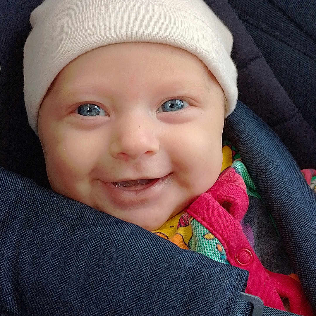 Alva is registered to the contest to win money with this photo: apparel, baby, blue_eyes, car_seat, child, closeup, colorful_clothing, cute, fabric, face, happy, hat, headwear, infant, portrait, safety_belt, sitting, smiling, strap, young_child
