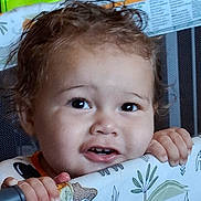 Lucius is registered to the contest to win money with this photo: toddler, child, baby, face, portrait, curly_hair, brown_hair, eyes, smile, teeth, hands, sitting, playpen, indoor, newspaper, toy, patterned_blanket, close_up, curious, happy