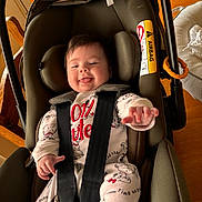 Lyra is registered to the contest to win money with this photo: baby, car_seat, infant, pajamas, smiling, indoor, child, seat_belt, cute, playful, tongue_out, wooden_furniture, table, chair, fabric, face, person, sitting, safety_harness, expression