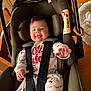 baby, car_seat, infant, pajamas, smiling, indoor, child, seat_belt, cute, playful, tongue_out, wooden_furniture, table, chair, fabric, face, person, sitting, safety_harness, expression