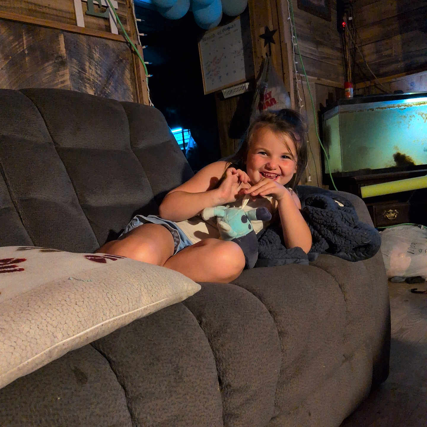 Kaydence is registered to the contest to win money with this photo: child, girl, smiling, couch, plush_toy, indoor, wooden_wall, balloons, fish_tank, blanket, pillow, casual_clothing, happy, living_room, relaxing, home, furniture, cozy, portrait, person