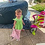 Arya participe au concours pour gagner de l'argent avec cette photo : toddler, child, green_dress, pink_blanket, barefoot, tricycle, outdoor, patio, hot_tub, plastic_toy, sunlight, footwear, fence, grass, concrete, shoe, person, playtime, daylight, casual