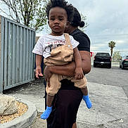 Kordae joined the competition — help win amazing prizes! toddler, child, adult, outdoor, urban, parking_lot, tree, metal_fence, clothing, overalls, tshirt, socks, person, holding, side_view, vehicle, asphalt, rock, sky, casual