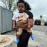 toddler, child, adult, outdoor, urban, parking_lot, tree, metal_fence, clothing, overalls, tshirt, socks, person, holding, side_view, vehicle, asphalt, rock, sky, casual