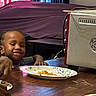child, smiling, table, paper_plate, food, indoor, curtains, appliance, metal, wood, person, dining, meal, kitchen, happy, face, hand, colorful, background, young
