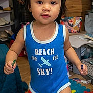 Cataleya Nykishia is registered to the contest to win money with this photo: toddler, child, indoor, blue_onesie, text_on_clothing, cap, crayons, colorful_playmat, floor, furniture, toy, person, young_child, standing, expression, hair, face, room, casual_clothing, playtime