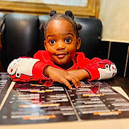 Hope is registered to the contest to win money with this photo: child, smiling, table, menu, restaurant, red_clothing, face, hair_buns, indoor, curious, person, cute, young, seated, hands, black_background, wooden_frame, casual, portrait, waiting