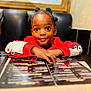 child, smiling, table, menu, restaurant, red_clothing, face, hair_buns, indoor, curious, person, cute, young, seated, hands, black_background, wooden_frame, casual, portrait, waiting