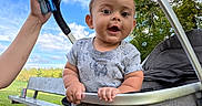 Kai joined the competition — help win amazing prizes! baby, child, stroller, outdoor, park, smiling, surprised, sitting, infant, portrait, face, person, adult, hand, trees, sky, grass, bench, handle, clothing