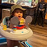 Kai is registered to the contest to win money with this photo: baby, child, infant, portrait, sitting, activity_center, toy, explorer_hat, mickey_mouse, batman, aquarium, fish_tank, indoor, playroom, furniture, wood_floor, smiling, surprised, colorful, family_home