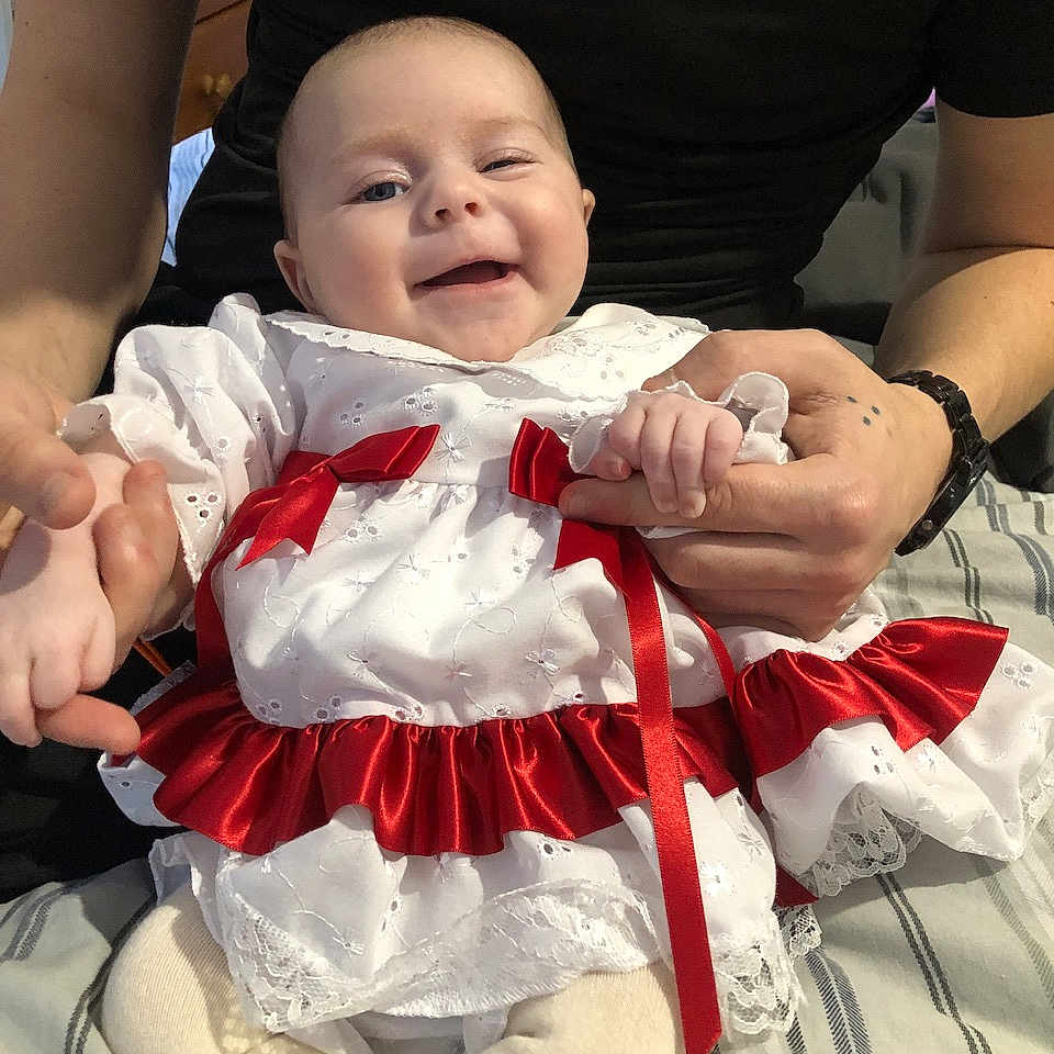 Olivia-May is registered to the contest to win money with this photo: adult_hands, baby, bed, black_shirt, child, cute, fabric, happy, holding_hands, indoors, infant, lace, person, portrait, red_ribbons, ribbons, skin, smiling, striped_bedspread, white_dress