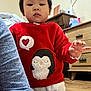 casual, child, clothing, cozy, dresser, expressionless, face, floor, furniture, hand, heart_symbol, home, indoor, long_sleeve, penguin_design, person, red_sweater, sleeve, toddler, wooden_floor