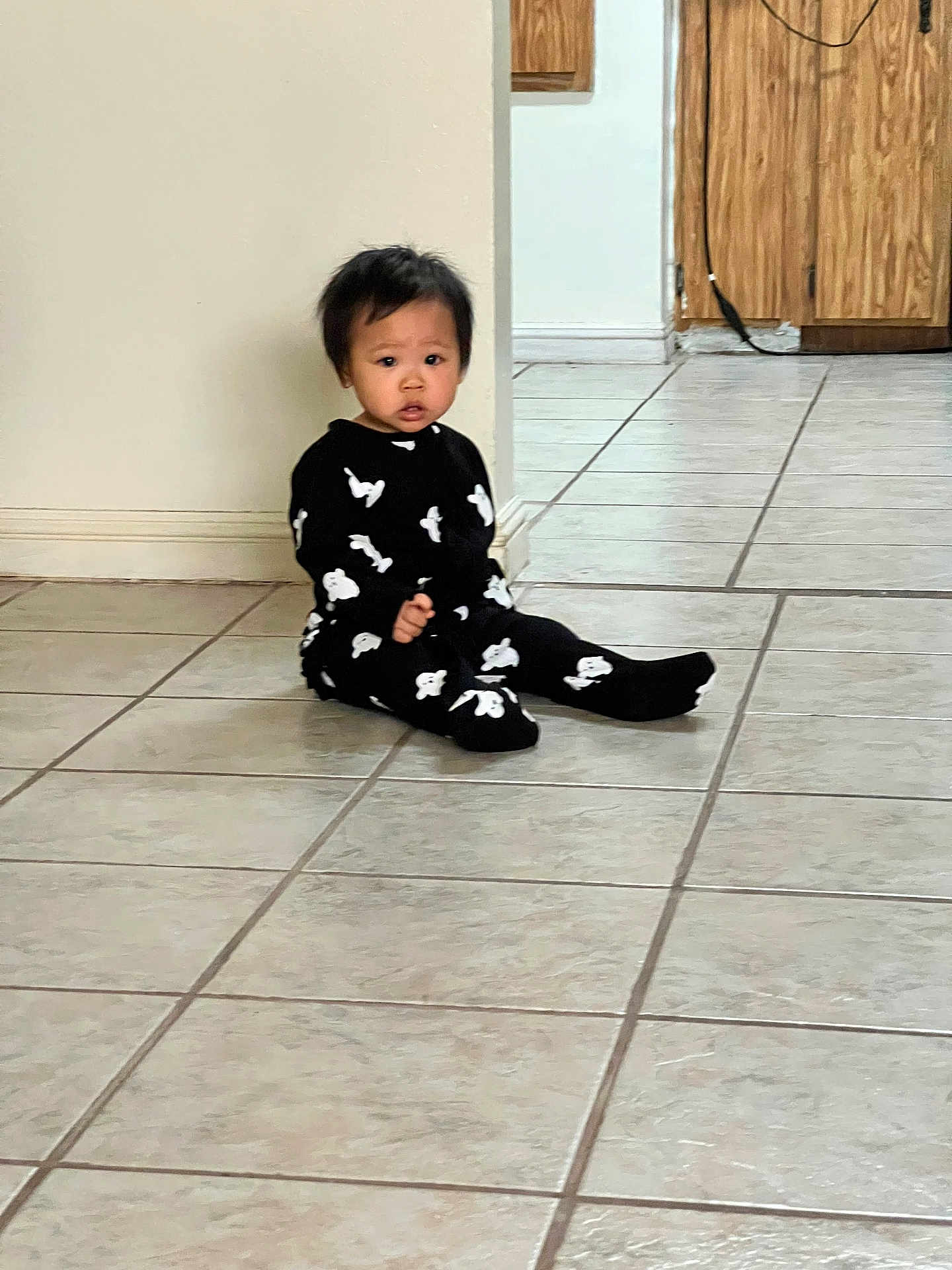 Tiffany joined the competition — help win amazing prizes! toddler, child, baby, onesie, black_clothing, white_pattern, sitting, floor, tile_floor, indoor, home, curious, expression, young_child, person, hair, face, footwear, cabinet, wall