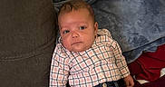 Armand is registered to the contest to win money with this photo: baby, child, infant, portrait, sitting, couch, blanket, plaid_shirt, jeans, pacifier, face, eyes, brown_hair, indoor, home, furniture, textile, red_cushion, blue_blanket, casual_clothing