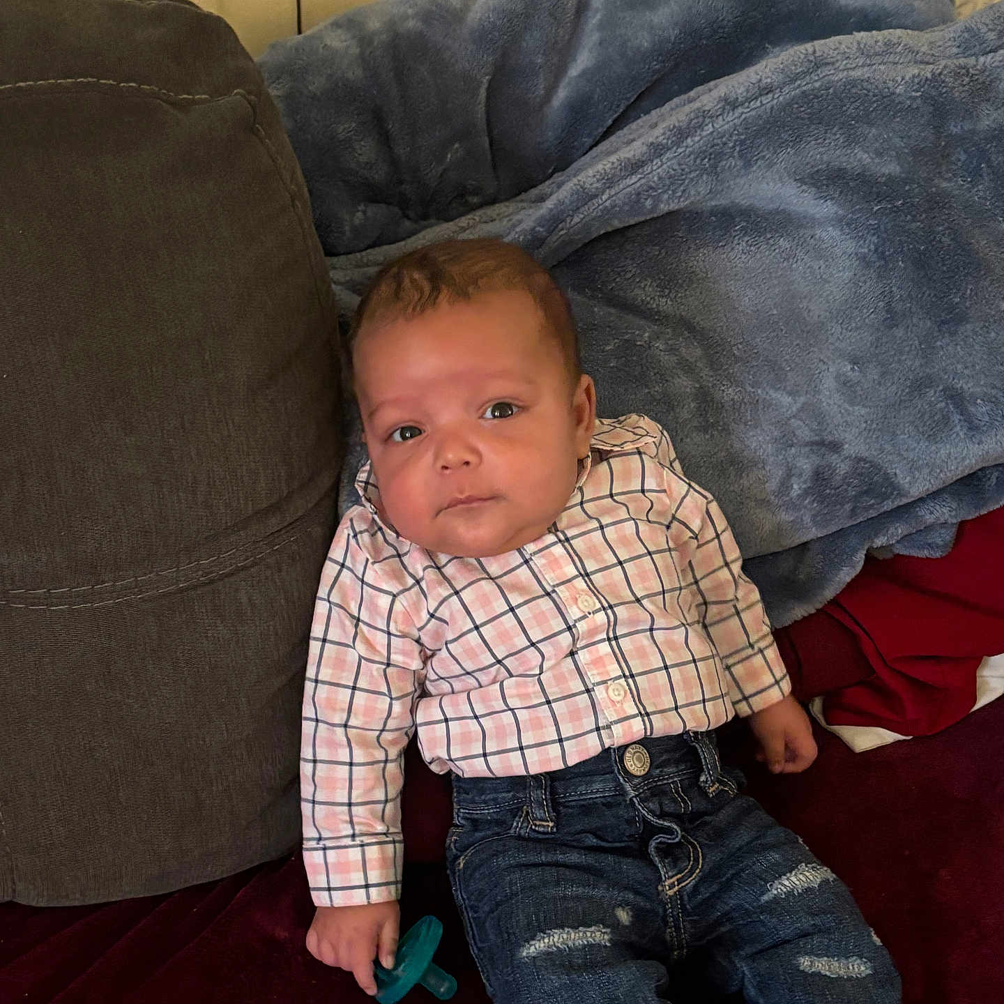 Armand is registered to the contest to win money with this photo: baby, child, infant, portrait, sitting, couch, blanket, plaid_shirt, jeans, pacifier, face, eyes, brown_hair, indoor, home, furniture, textile, red_cushion, blue_blanket, casual_clothing