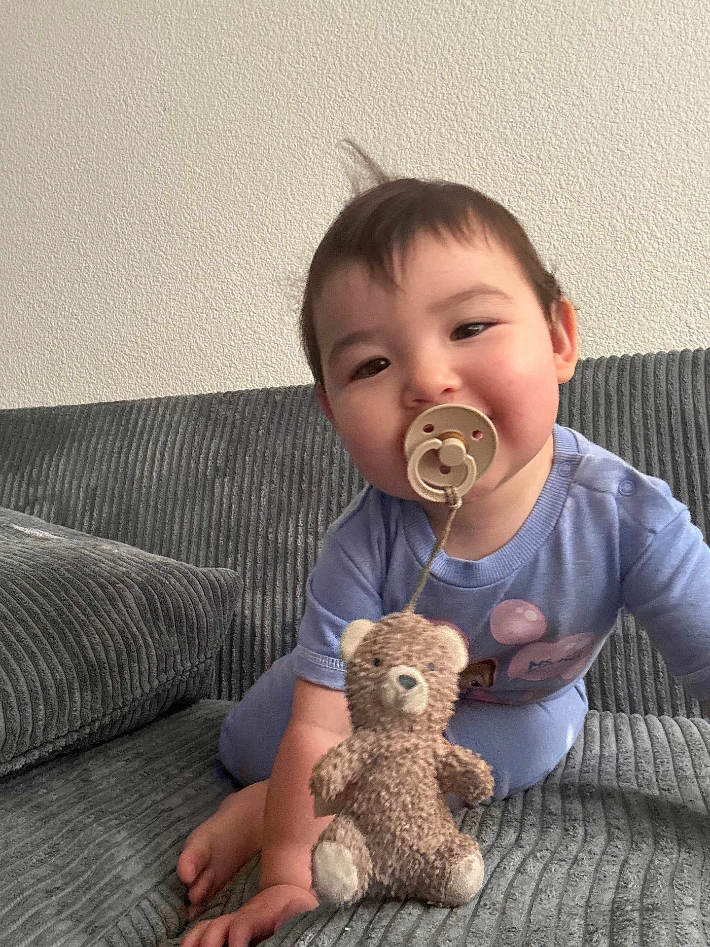 Amaia is registered to the contest to win money with this photo: baby, pacifier, teddy_bear, plush_toy, couch, grey_couch, child, indoor, cute, infant, smiling, blue_clothing, toddler, hair, face, sitting, small_toy, home, comfort, young_child