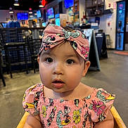 Amaia is registered to the contest to win money with this photo: baby, child, pink_headband, minnie_mouse, dress, high_chair, restaurant, indoor, curious, face, food_on_lips, colorful, lights, television, furniture, wooden_chair, person, cute, portrait, casual