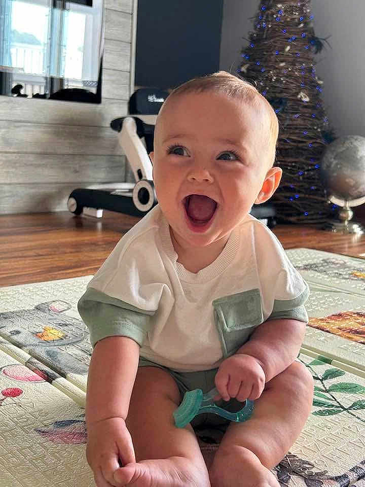 Bennett is registered to the contest to win money with this photo: baby, child, smiling, sitting, teething_toy, play_mat, indoor, living_room, christmas_tree, holiday_decor, treadmill, wood_floor, happy, cute, infant, portrait, short_sleeve_shirt, expression, toy, home