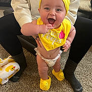 Bennett joined the competition — help win amazing prizes! baby, child, smiling, yellow_hat, yellow_bib, diaper, indoor, carpet, adult_hands, baby_slippers, yellow_slippers, person, happy, cute, infant, toddler, clothing, home, holding, support