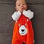 baby, infant, child, onesie, orange_clothing, bear_design, hand, face, expression, lying_down, curious, cute, sucking_fingers, white_sleeves, fabric, soft_surface, indoor, portrait, young_child, playful