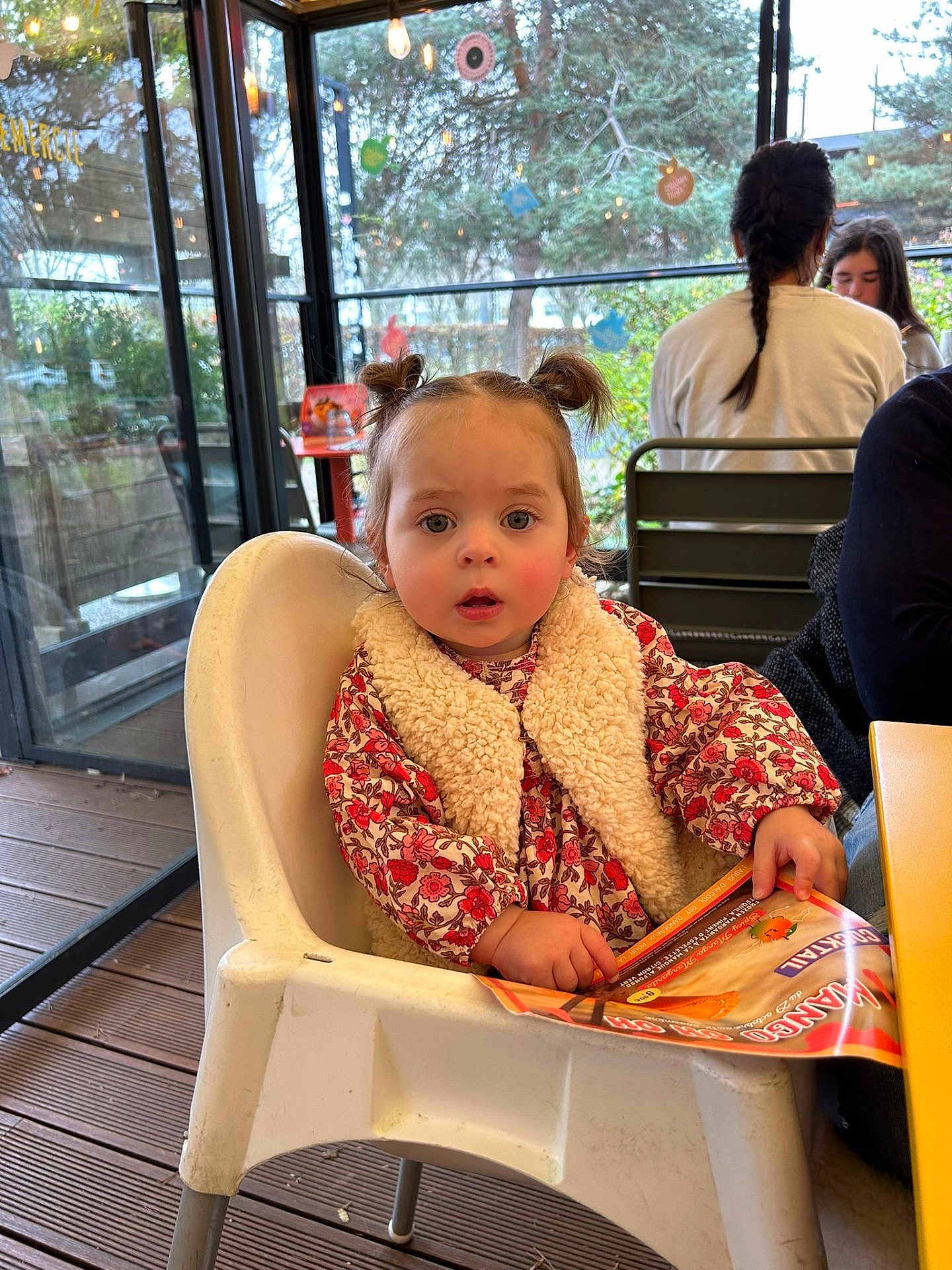 Alba participe au concours pour gagner de l'argent avec cette photo : toddler, child, high_chair, menu, pigtails, indoor, cafe, glass_wall, table, chair, person, red_flower_pattern, vest, wood_floor, people, braid, casual_clothing, window, daylight, expression