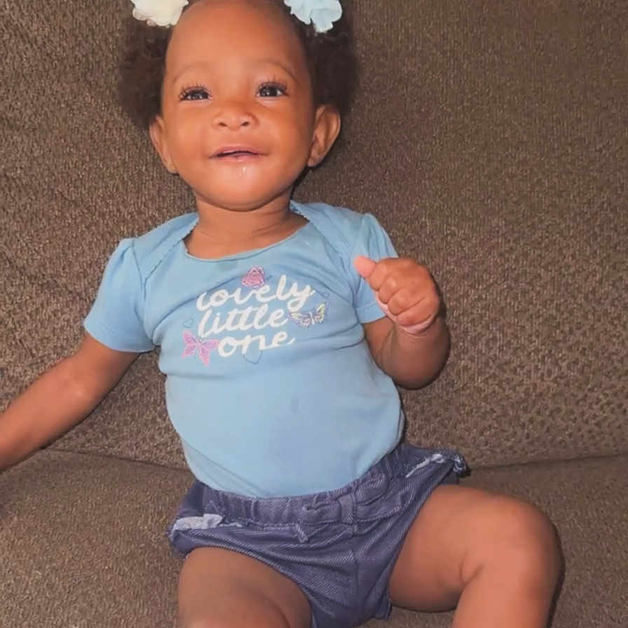 Unique is registered to the contest to win money with this photo: baby, blue_shirt, child, clothing, couch, cozy, cute, face, flower_hair_clips, happy, home, indoor, infant, person, pink_slippers, portrait, sitting, skin, smiling, young_child