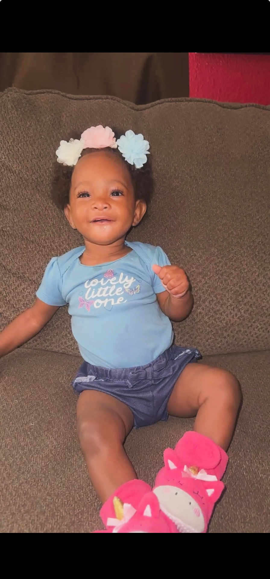 Unique is registered to the contest to win money with this photo: baby, child, smiling, blue_shirt, pink_slippers, flower_hair_clips, couch, indoor, cute, happy, portrait, sitting, infant, clothing, person, young_child, home, cozy, skin, face