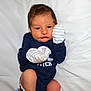 baby, bodypart, clothing, cushion, face, finger, glove, hand, happy, head, homedecor, indoors, newborn, person, photography, portrait, shirt, sitting, smile, t_shirt