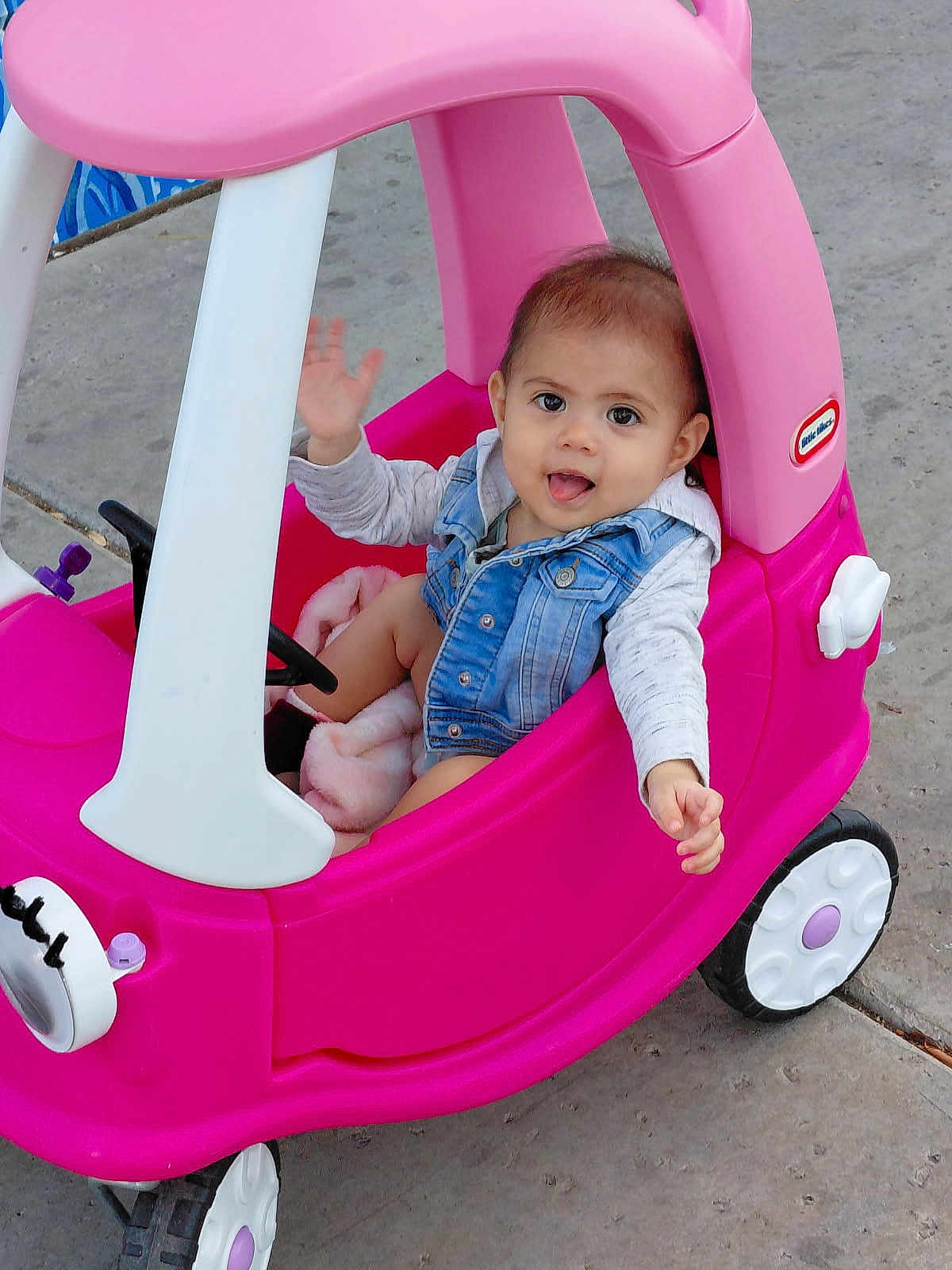 Nita is registered to the contest to win money with this photo: baby, child, toddler, toy_car, pink_car, little_tikes, denim_jacket, blanket, waving, tongue_out, smile, steering_wheel, wheel, plastic_toy, outdoor, concrete, sidewalk, portrait, playtime, cute
