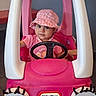 child, clothing, cute, expression, eyes, face, floor, hat, indoors, person, pink, plastic, playful, seat, small, smiling, steering_wheel, toddler, toy_car, vehicle