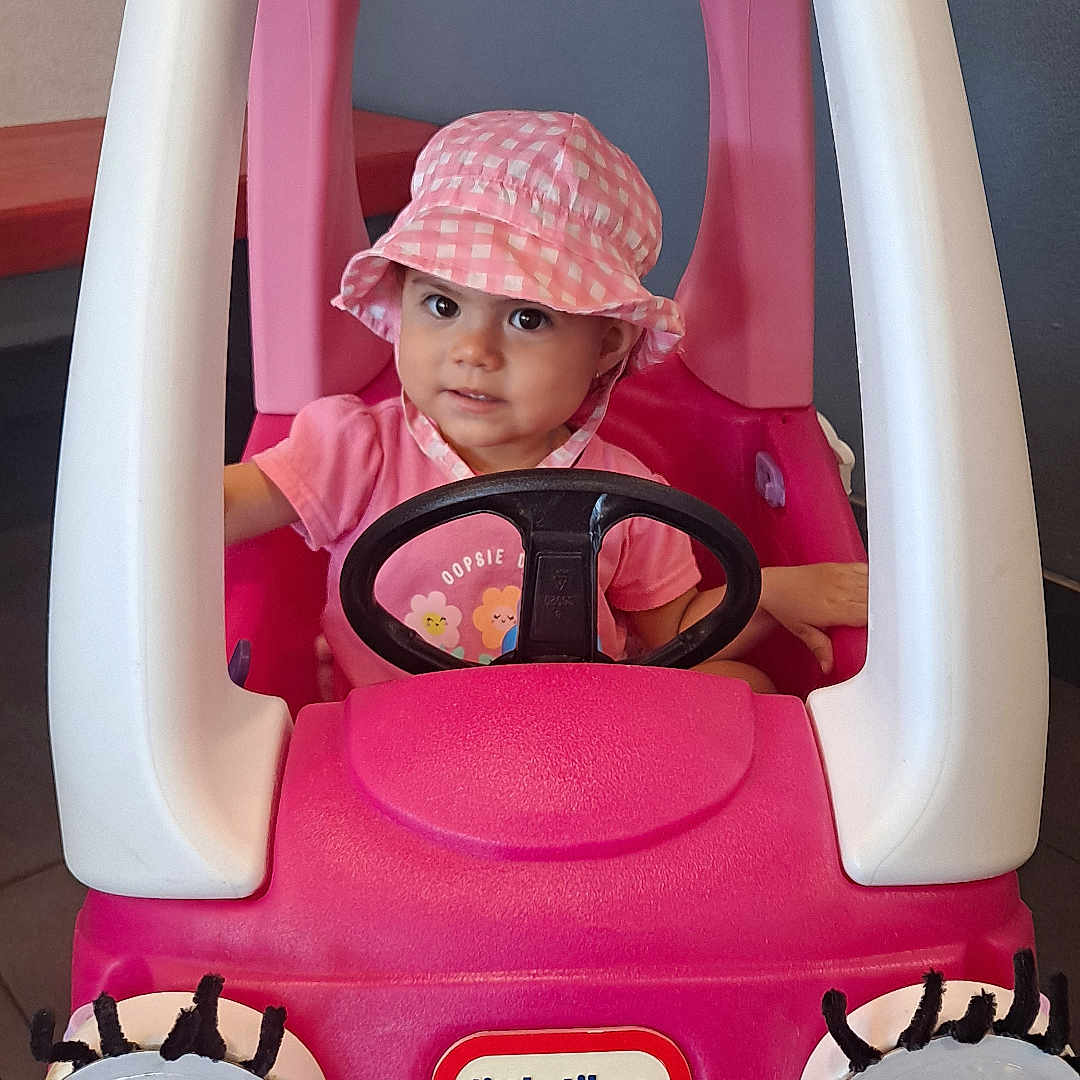 Nita joined the competition — help win amazing prizes! child, clothing, cute, expression, eyes, face, floor, hat, indoors, person, pink, plastic, playful, seat, small, smiling, steering_wheel, toddler, toy_car, vehicle