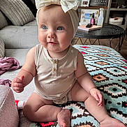 Wrenlee is registered to the contest to win money with this photo: baby, child, infant, headband, bow, blue_eyes, sitting, blanket, patterned_blanket, living_room, couch, furniture, wood_floor, wooden_table, bottle, clothing, cute, indoors, person, smiling