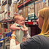 aisle, baby, blond_hair, boxes, clothing, flooring, happy, holding, infant, interaction, joy, person, retail, shelves, shopping, shopping_cart, smiling, store, warehouse, woman