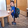 child, backpack, blue_shirt, shorts, sneakers, toy_car, table, indoor, floor, door, wall, cup, smiling, playful, person, standing, happy, furniture, casual, young