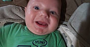 Janson is registered to the contest to win money with this photo: baby, child, infant, green_shirt, dinosaur_graphic, pacifier, bed, blanket, happy, smiling, face, person, indoors, toddler, clothing, cute, head, mouth_open, lying_down, young
