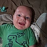 baby, child, infant, green_shirt, dinosaur_graphic, pacifier, bed, blanket, happy, smiling, face, person, indoors, toddler, clothing, cute, head, mouth_open, lying_down, young