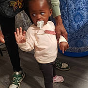 Maleah joined the competition — help win amazing prizes! toddler, child, pacifier, person, adult_hand, sneakers, floor, curtain, indoor, clothing, standing, pink, black_hair, pattern, footwear, holding, looking_away, long_sleeve, pants, home