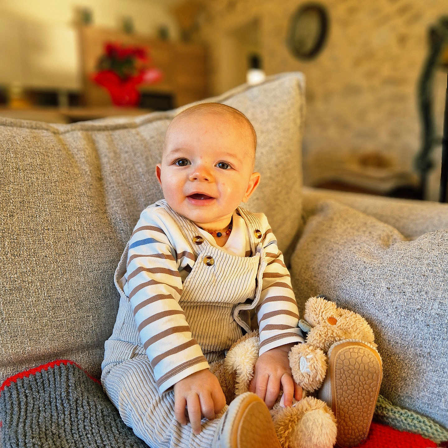 Lisandre a rejoint le concours — aidez-le/la à gagner de superbes lots ! baby, blurred_background, child, corduroy_overalls, couch, cozy, decor, face, furniture, happy, home_interior, indoor, knitted_blanket, person, plush_toy, seated, smiling, striped_clothing, teddy_bear, wood_beam