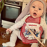 Giulia participe au concours pour gagner de l'argent avec cette photo : baby, child, apron, kitchen, whisk, wooden_spoon, bowl, cooking, cooking_utensils, face, expression, sitting, table, clothing, headwear, cute, indoors, person, light, gingham