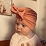 baby, child, headwrap, orange, clothing, hand, indoor, portrait, cute, infant, person, face, expression, wall, framed_picture, soft_lighting, skin, sitting, cozy, warm_tones