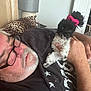 beard, black_and_white_dog, casual_clothing, companion, cozy, curious, dog, glasses, hair_tie, hand, holding, home, indoor, leopard_print, lying_down, man, pet, pillow, relaxed, short_hair