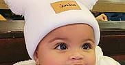 Maia is registered to the contest to win money with this photo: baby, child, infant, portrait, face, eyes, looking_away, curious, white_hat, pom_pom_hat, winter_clothing, puffy_jacket, earrings, seated, indoor, restaurant, cafe, background, close_up, photography