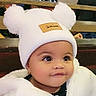 baby, child, infant, portrait, face, eyes, looking_away, curious, white_hat, pom_pom_hat, winter_clothing, puffy_jacket, earrings, seated, indoor, restaurant, cafe, background, close_up, photography