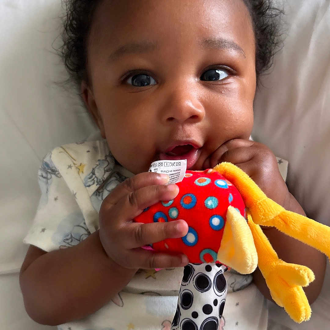 Jnari joined the competition — help win amazing prizes! baby, infant, child, toy, plush_toy, bed, onesie, cute, curly_hair, hand, face, chewing, lying_down, white_bed_sheet, colorful, patterned_clothing, expression, indoor, soft_toy, young_child