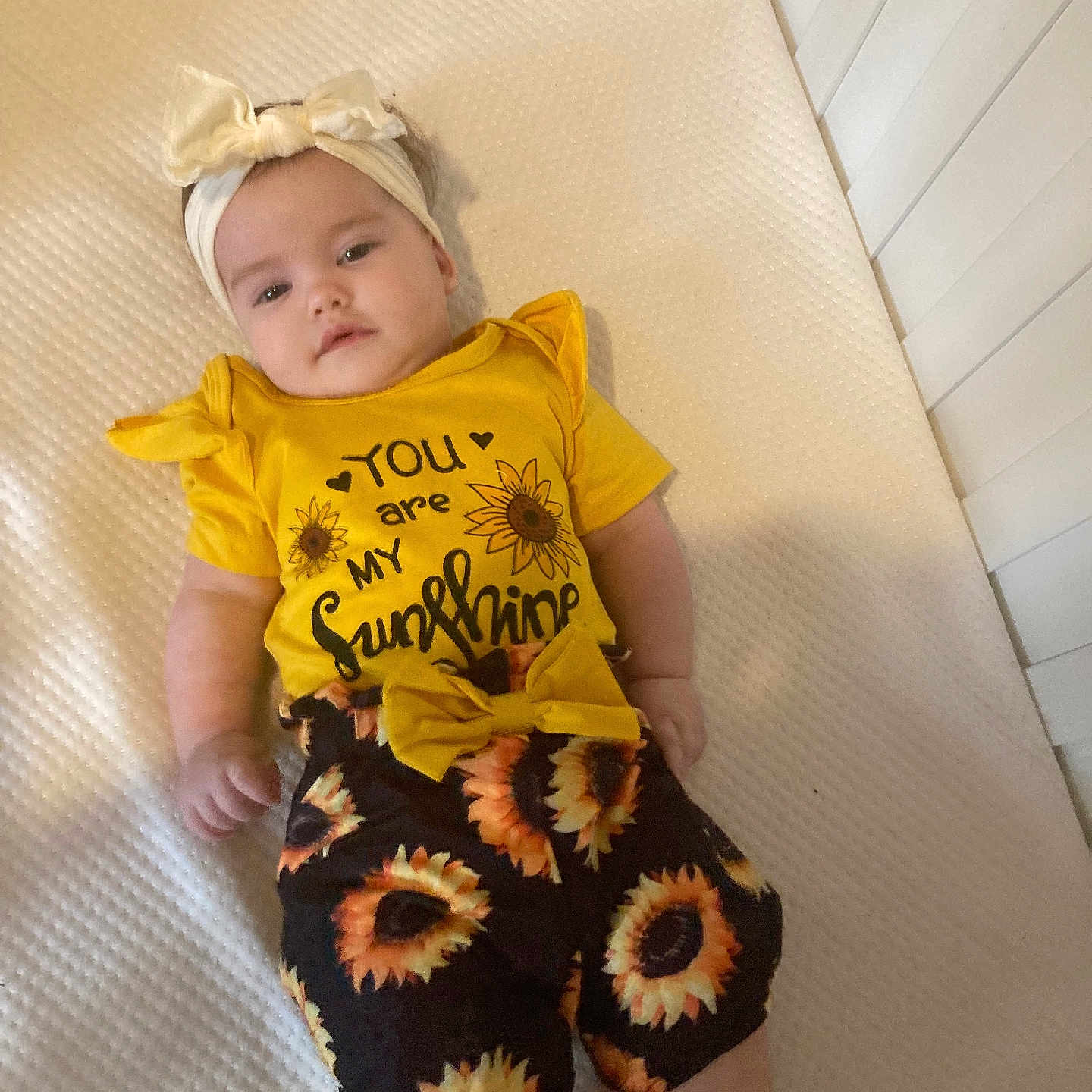Kensleigh joined the competition — help win amazing prizes! apparel, baby, bow, child, crib, cute, headband, indoors, infant, lying_down, mattress, playpen, portrait, smiling, soft_lighting, sunflower_pattern, text_on_clothing, toddler, white_background, yellow_clothing